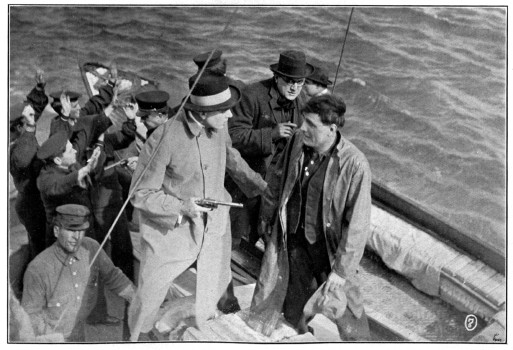 Several men stand in a boat with their hands up, with guns pointing at them.
