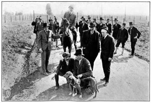 Several men stand around a pair of men crouched next to two bloodhounds.