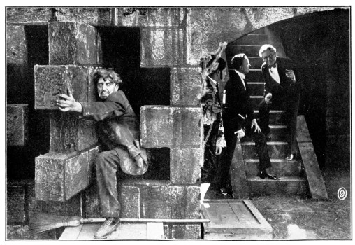 A group of men congregate on stone stairs, while a man desperately grasps a secret door in the stone wall.