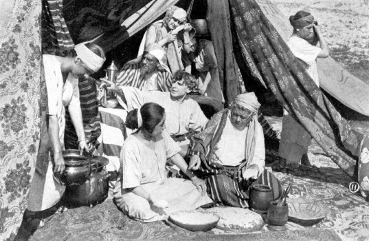 A group sits outside a tent in the desert, while a man gesticulates.