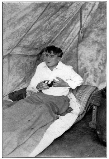 A man sits up on a cot in a tent, holding a small black box.