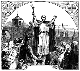 A man stands on a raised platform in the midst of a crowd. His arms are raised and his right hand holds a large cross.